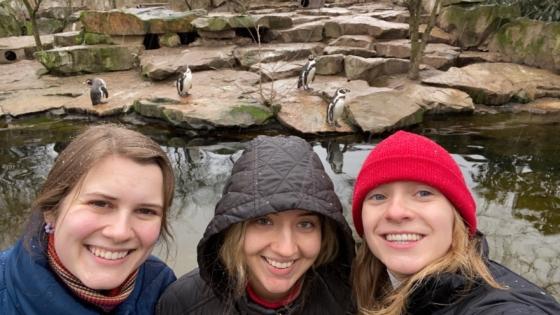 three young women stand in front of a zoo filled with penguins