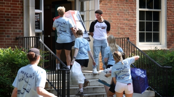 a group of young people carry dorm supplies into a dorm on a sunny day while wearing matching blue tshirts