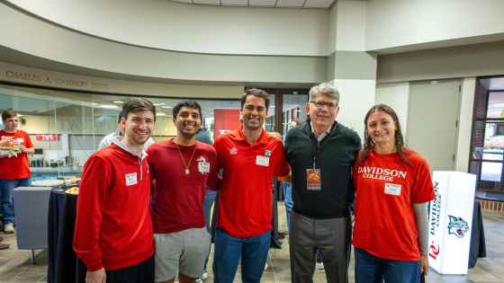 Five people pose for a photo at a Davidson College event near the Charles A. Cannon Pool, with four individuals wearing red college apparel and name tags alongside an older man in a black quarter-zip sweater.