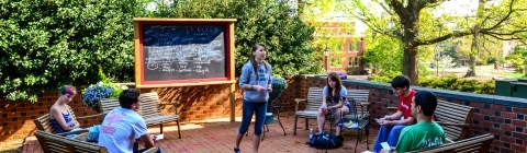 Russian Studies AT Session in outdoor classroom where students sit on benches and instructor addresses them in front a chalkboard with writing in Russian