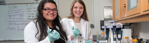 Two students stand in neuroscience lab holding pipettes and research equipment