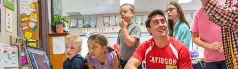 Davidson student wearing a Davidson soccer jersey stands amidst a group of young kids in their classroom and shows them math exercises on a laptop