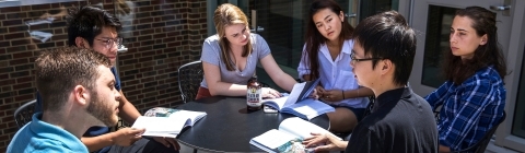 Group of students sits around a round table holding books and talking while on a patio