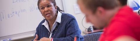 Professor Brenda Flanagan sits with students in class and gives lecture while students take notes