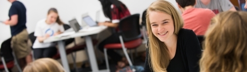 Student talks to classmates sitting at same table. Rest of class is out of focus in the background