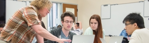 Professor Heyer stands near table and talks to three students who are using laptops in her class