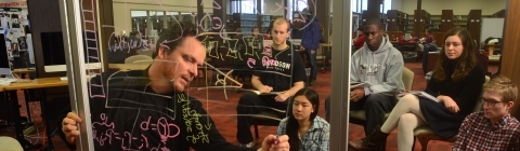 Professor Tim Chartier writes on a glass white board while leading a class to a group of students in the library