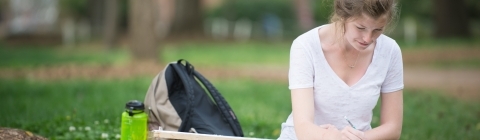 Student sits on grass with oversized drawing pad while drawing her campus surroundings