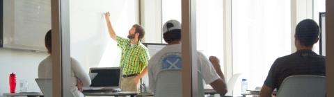 Professor writes on board as students backs are shown looking at him through glass classroom walls