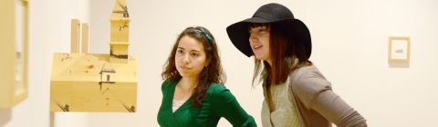 Two students stand near a work that is shaped like a house and is 3d, sticking out of the wall