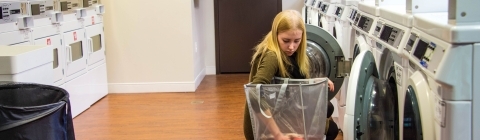 Student kneels next to clothes hamper while doing laundry