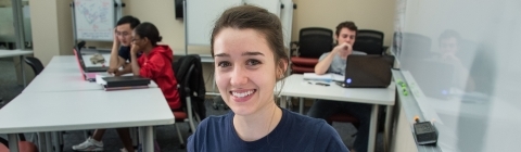 Student smiles at camera with tutoring supplies in front of her and people being tutored in the background