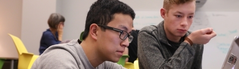 Students sit at table and while one types on keyboard, other student watches