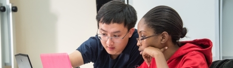 Two students talk with laptops open, one pointing at his colleague's laptop