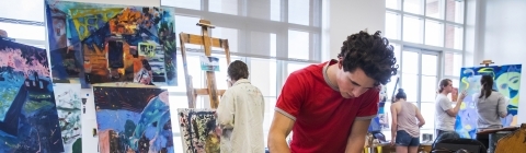 Student mixes paint in foreground while other students paint canvases in background