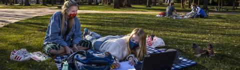 Two Friends Sitting on Blanket on Lawn with Masks on Laughing and Studying