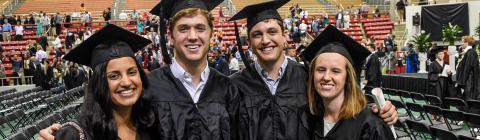 Students smile in caps and gowns at indoor Commencement