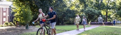 Two students walking, one holding a bike and talking on campus