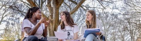 Three students sit on a picnic bench and converse over books