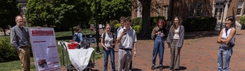 A group of students and faculty stand around and smile as a student drives a go-kart