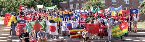 A large group of students in front of a brick building holding flags from around the world