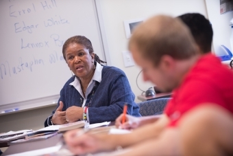 Professor Brenda Flanagan sits with students in class and gives lecture while students take notes