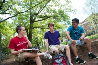 Three students sit on a brick wall, one holding a textbook and discussing