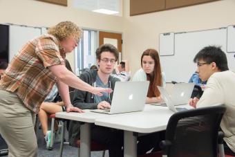 Professor Heyer stands near table and talks to three students who are using laptops in her class