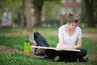 Student sits on grass with oversized drawing pad while drawing her campus surroundings