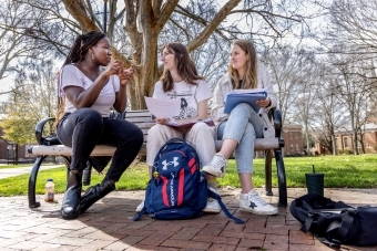 Three students sit on a picnic bench and converse over books