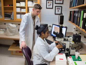 Two students in lab coats conduct research with scientific equipment including a microscope