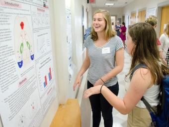 Student presents on her research standing by a poster and talking to a fellow student