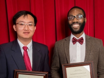 Two students hold framed awards in front of velvet red curtain