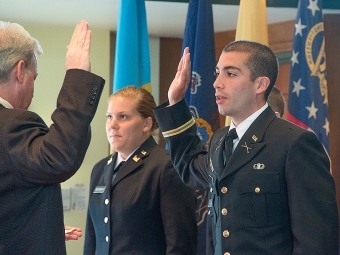 Two students stand in front of a row of flags, one has hand in the air mimicking an ROTC leader standing in front of him