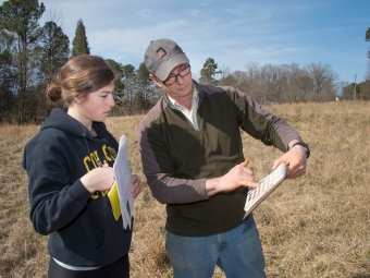 Prof. Dave Backus points to chart on clipboard in a field, student listens to what he is pointing out