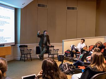 Professor Denham stands near projector screen, leaning on chair, at the front of auditorium while students, in seats, listen