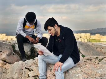 Two students sit on marble blocks at the Acropolis, one holding a reading he is reciting