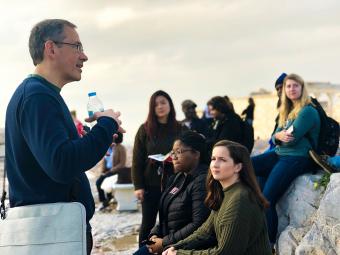 Prof. Randy Ingram speaks to class while holding a water bottle as students sit on a bench and on marble blocks at the Acropolis