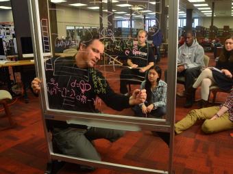 Professor Tim Chartier writes on a glass white board while leading a class to a group of students in the library