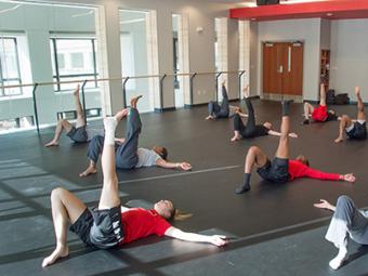 A large group of students in dance class lay on mats in the dance studio and hold their legs in the air