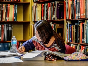 Student Studying in Library sitting at table with textbooks and surrounded by bookshelves