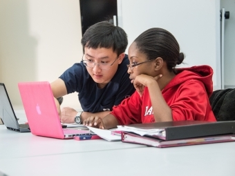 Two students talk with laptops open, one pointing at his colleague's laptop