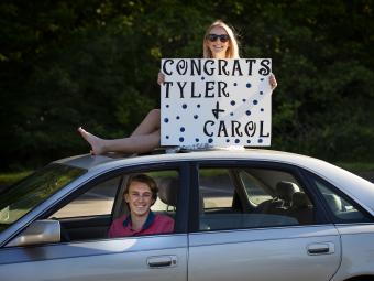 Friend sitting on top of car with sign "Congrats Tyler & Carol" 