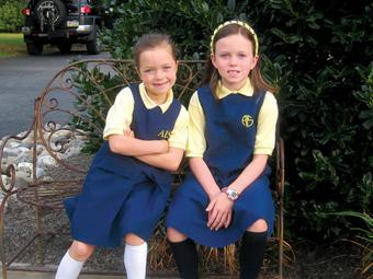  Olivia and Julianne Carey as children in school uniforms sitting on a bench outside
