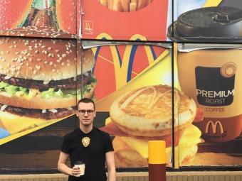 Davidson College alum Dan Van Note '14 with cup of cuffee in front of large McDonalds fast food street billboard