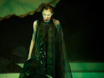 Emma acting on stage with black costume, red fingerprint on her face, and dark green glow on stage 