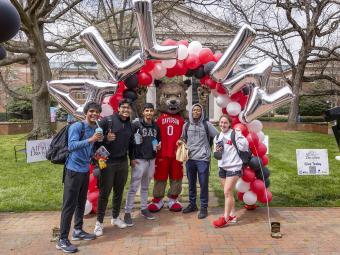 group of students smiling with mascot Lux the Wildcat in front of balloon garland with letters "All In"  