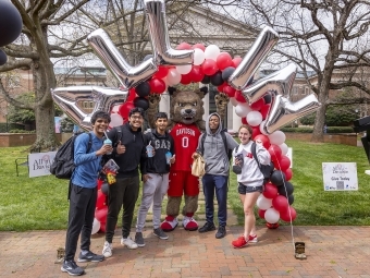 group of students smiling with mascot Lux the Wildcat in front of balloon garland with letters "All In"