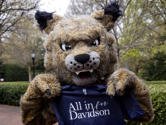 Mascot Lux the wildcat holds t-shirt that says "All in for Davidson" outside on campus