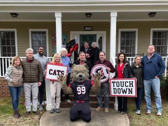 Peter Andrews, group of Davidson staff including president & athletic director, and Lux the wildcat outside Dave & Barbara Fagg's house with Davidson signs  "Cats" and "Touch Down" in hands